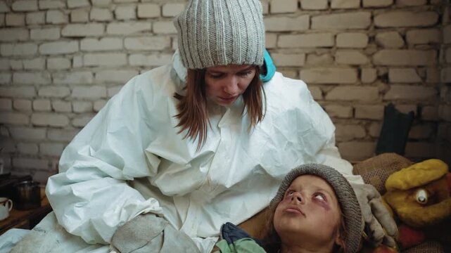 Rescuer in white hazmat suit and knitted hat kneels beside abandoned girl with bruised face and dirty clothing, lying on makeshift bedding, looking up with vulnerable eyes in cold brick shelter