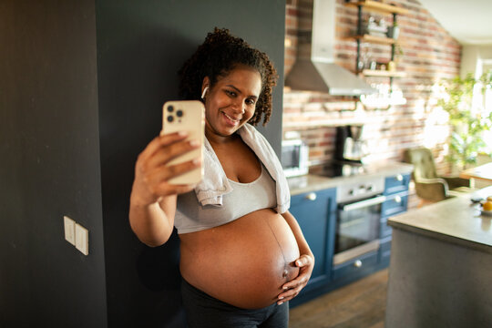 Pregnant woman taking belly selfie in home kitchen