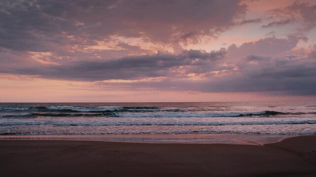 Beautiful sunrise over gentle sea waves rolling onto a sandy beach under soft pastel clouds during dawn.