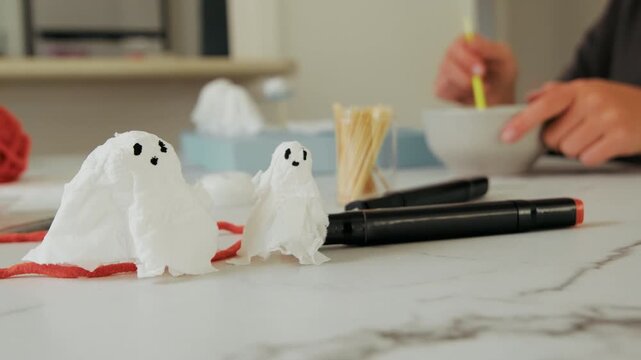 Small ghost decorations crafted from white tissues and markers resting on a table during a fun indoor holiday activity, person crafting handmade ghost figures using paper tissues and ink pens