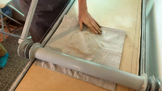 Woman rolls clay slab through roller between canvas sheets
