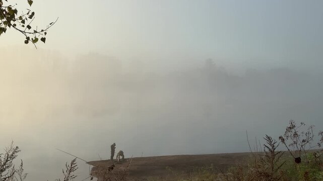 Two fishermen on a riverbank in dense autumn morning fog
