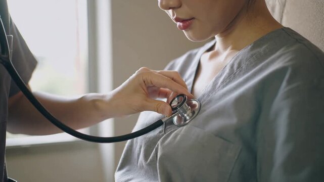 Approaching clinician placing stethoscope on woman in grey scrubs in exam room, checking heartbeat