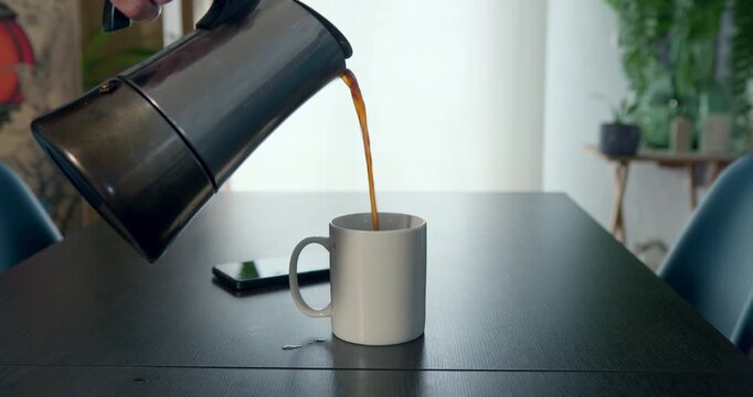 Brewing Morning Ritual: A steaming cup of coffee is poured from a moka pot into a white mug, set on a dark wooden table, hinting at a quiet morning routine and the start of a new day.