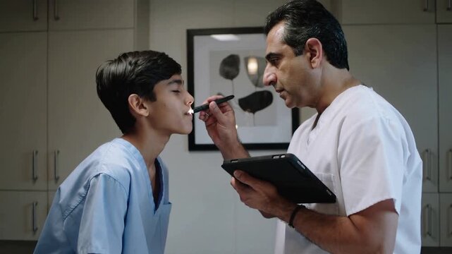 Inspecting clinician in scrubs leaning over teen after opening mouth in clinic, using tablet, light
