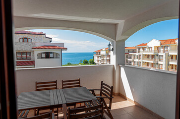 Fototapeta premium A sunny balcony with wooden furniture offers a scenic view of coastal buildings and the blue sea.