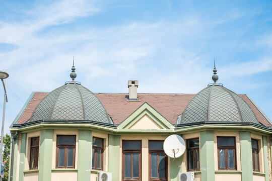 Ornate building facade with twin domed slate roofs, decorative spires, tiled gable roof, tall windows, satellite dish, and blue sky