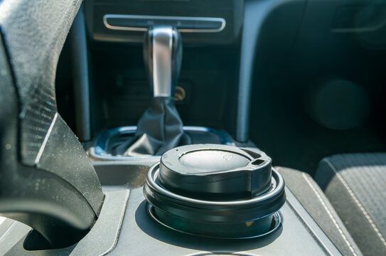 Black takeaway coffee cup with lid in a car cup holder beside the gear shift, surrounded by the vehicle center console and seats