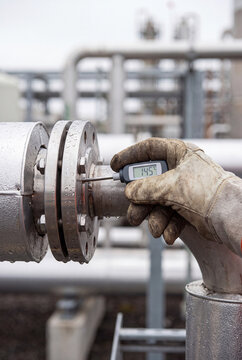 A gloved worker holding a digital thermometer measuring the high temperature on a industrial pipe flange.