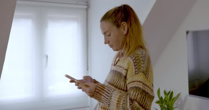 A woman shopping on her mobile from the comfort of her own home: A 50-year-old woman with her hair tied back in a ponytail and wearing a printed T-shirt is focused on her smartphone.