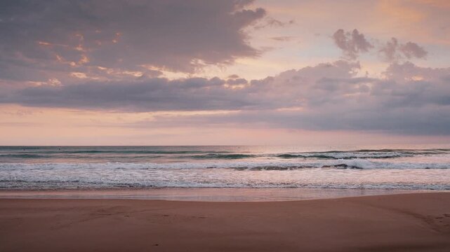 Beautiful sunrise over gentle sea waves rolling onto a sandy beach under soft pastel clouds during dawn.