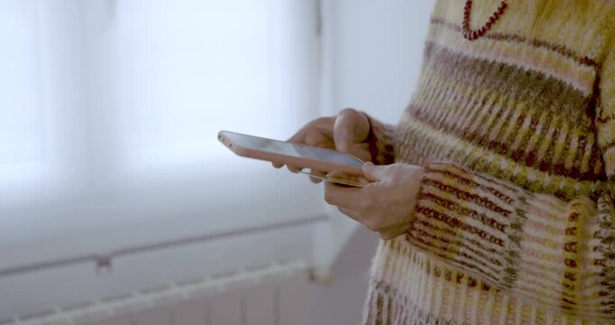 Holding a card and a smartphone: A person&rsquo;s hands, clad in a knitted jumper, gently hold and operate a modern smartphone by a window.