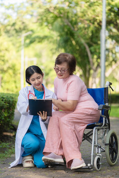 Asian female doctor holding clipboard and discussing medical results with senior woman in wheelchair in park