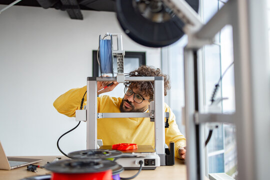 An Indian man with curly hair and glasses is carefully lubricating the axles of a 3D printer during routine maintenance in his workshop