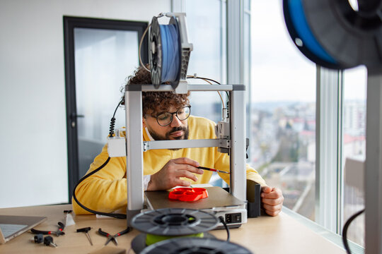 An Indian man with curly hair and glasses performs routine maintenance on a 3D printer, lubricating its axles with a small brush