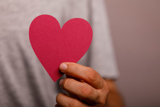 A person's hand gently holds a bright red paper heart, symbolizing love and compassion.