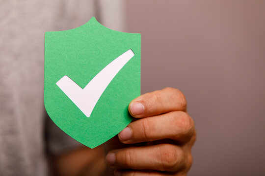 Hand holding a green paper shield with a white checkmark, symbolizing approval or safety.