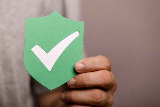 A hand holds a green paper shield with a white checkmark, symbolizing safety and verification.