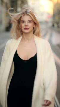 Slow motion 120fps vertical video of blonde woman running on European street and turning to camera with flying hair. Beautiful female model in white cardigan strolling outdoors in evening light