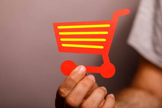 A person's hand holds a vibrant red paper shopping cart, symbolizing online retail and consumerism.