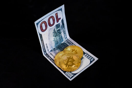 Three physical golden Bitcoin token coins arranged overlapping on top of a single folded United States 100 dollar banknote.