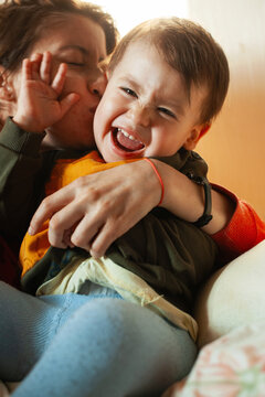 Mother kissing laughing toddler in warm close-up family hug, playful bonding as child squirms in comfy casual clothing under bright indoor light, affectionate
