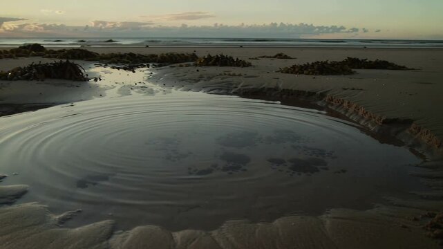 Rippling tidal pool sending expanding rings across low-tide beach at sunrise, triggered by impact