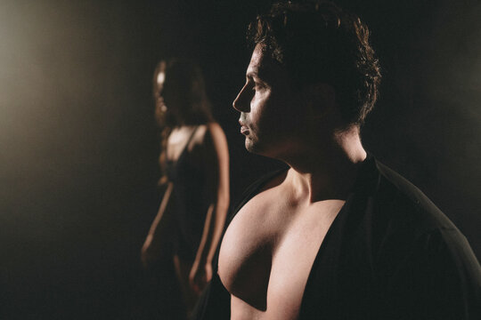 Shirtless young man with partially open dark jacket in dramatic side lighting, muscular torso foreground, blurred woman in dress in shadowy background