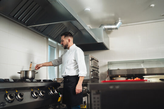 Male chef stirring saucepan on commercial stove in professional kitchen with stainless steel appliances and ventilation hood