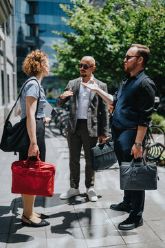 Three business colleagues talk and gesture while holding briefcases on a city sidewalk. A candid urban office conversation between professionals during a sunny workday outdoors.