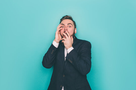 Horizontal picture of young man. He is scared and terrified. Guy has closed the part of his face with his hands. Isolated on blue background.