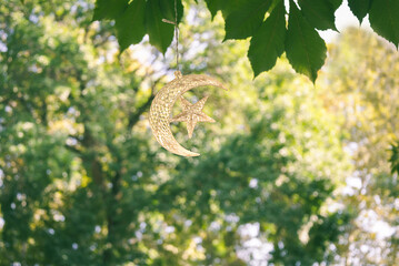 White crescent moon and star hanging decoration against a softly blurred green garden background. © Irina