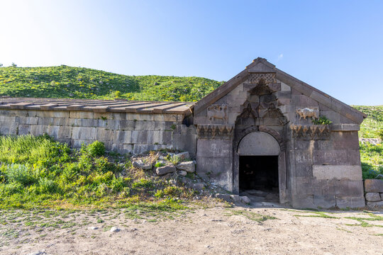 Basalt gabled entrance of the Orbelian Caravanserai with carved ram and bull reliefs above a dark arched doorway and a stone roof beside a green grassy hillside, Selim, Armenia