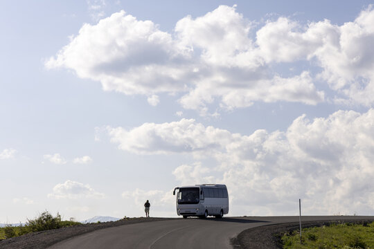Silhouette of a lone traveler walking toward a white tour bus on a winding mountain road beneath huge white clouds in a soft blue sky near Selim Pass, Selim, Armenia