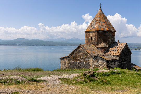 Sevanavank Monastery church with a conical orange stone roof on a grassy hilltop above the calm blue water of Lake Sevan with distant mountains and clouds, Sevanavank, Armenia