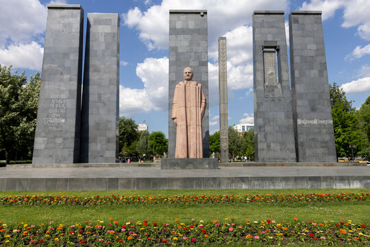 Pink granite statue of Alexander Myasnikyan standing between tall dark granite memorial slabs with Armenian inscriptions above a colorful flower bed, Yerevan, Armenia