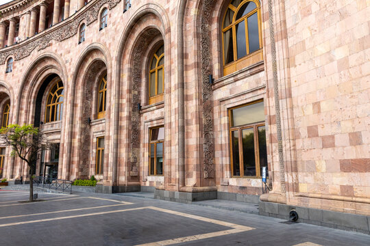 Ornate carved pink tufa stone arches with tall wooden framed windows and floral relief columns on a historic building facade beside an empty paved square, Yerevan, Armenia