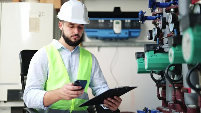 Diligent technician performing inspection, noting data on a smartphone and clipboard. Wearing a hard hat and safety vest, observing complex piping and gauges in a boiler room.