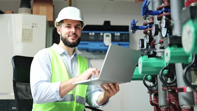 Confident engineer with a beard, wearing a white hard hat and high-visibility vest, inputs data on a laptop in an industrial boiler room, then smiles. Expert performs system check and maintenance.