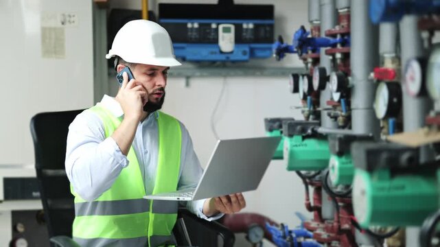 Focused engineer, wearing a white hard hat and high-visibility vest, communicates on a smartphone while reviewing data on a laptop within a complex industrial boiler room setting.