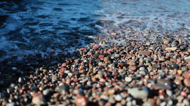 Waves rolling onto colorful pebble beach on seashore. Smooth sea stones close up with blue water surf in background. Nature coastal landscape of rocky beach