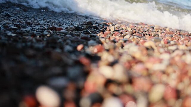 Sea wave washing colorful pebble beach. Close-up of foamy surf flowing over stones on seashore. Coastal nature background with shallow depth of field.
