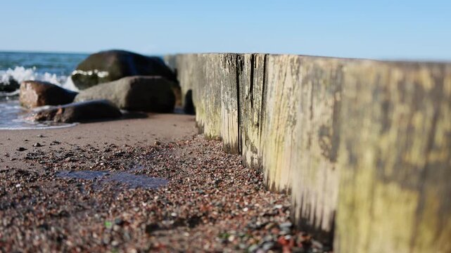 Old wooden breakwater on pebble beach. Sea waves washing stony shore with large rocks. Coastal protection groyne under blue sky. Baltic seaside nature.