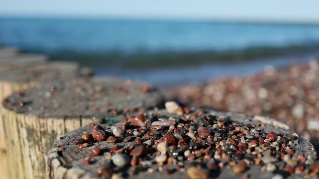 Small pebbles and stones lying on old wooden groins on pebble beach. Sea waves rolling on shore in blurred background. Coastal nature detail, calm seascape