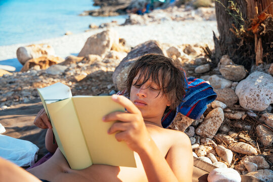 teenage boy reading a book while relaxing on a rocky beach
