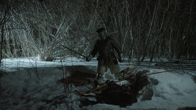 Armed medieval knight wearing chainmail and helmet stands over prone figure in snow at night holding weapon. Scene is set outdoors in snowy woodland with bare trees under dim lighting