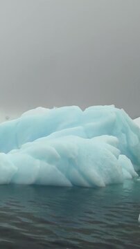 Vertical Video of Low Sculpted Icebergs Along the Horizon. White and blue ice, rippling water and soft diffuse light reveal natural frozen texture and sculpted contours across the marine scene.