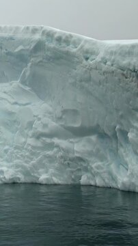 Vertical Video of Rounded White Iceberg Above Rippling Dark Water. White and blue ice, rippling water and soft diffuse light reveal frozen texture and sculpted contours.