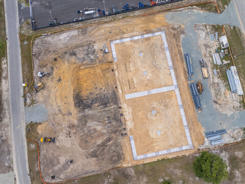 Aerial view of a construction site featuring concrete foundations, heavy machinery, and building materials on a dirt lot in Leland, North Carolina, United States.