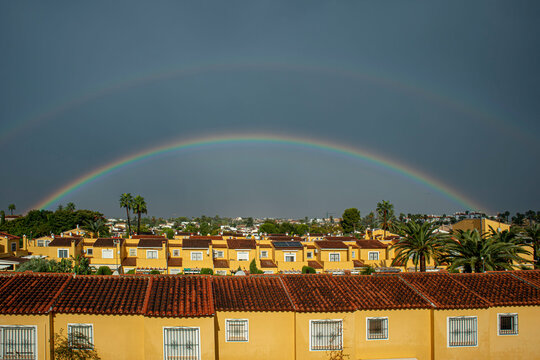 Vibrant double rainbow over yellow Mediterranean houses with stormy sky
